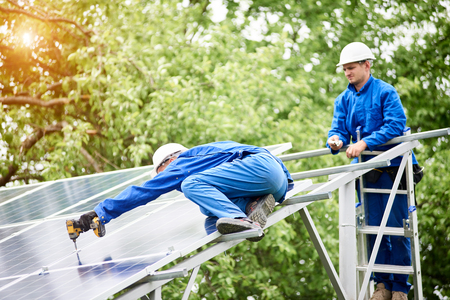 Team of two constructors connecting photo voltaic panel to stand-alone solar system platform using screwdriver. Alternative energy, professionally done job and profitable financial investment concept.の写真素材