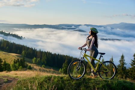 Sporty girl cyclist riding on yellow bicycle on a rural trail in the mountains, wearing helmet, enjoying valley view on sunny morning. Foggy mountains, forests on the blurred backgroundの写真素材