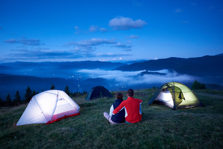 Back view, loving people sitting near camping enjoying sunrise in the mountains. In the valley between the mountains the lights of the town are burning which are covered with morning mountain hazeの写真素材