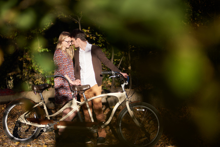 Happy romantic couple, bearded man and attractive woman close together at tandem double bicycle outdoors in dark autumn park or forest in the evening on blurred golden green bokeh background.の写真素材