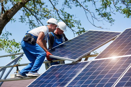 Two young technicians installing heavy solar photo voltaic panel on tall steel platform on green tree background. Exterior solar panel voltaic system installation, dangerous job concept.の写真素材
