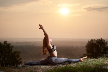 Profile of attractive slender long-haired barefooted young woman in summer training outfit doing gymnastic yoga exercises outdoors on grassy hill under bright pink misty sky at sunset or sunrise.の写真素材