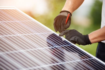 Close-up technicians hands in protective gloves making mark with pencil on metal platform for installing solar photo voltaic panel on blurred green summer background.