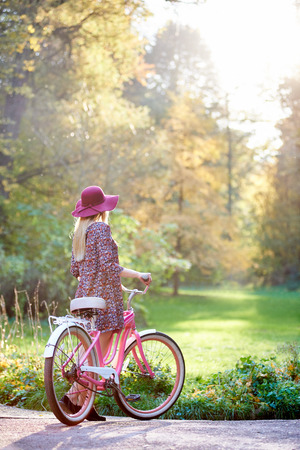 Back view of slim blond fashionable long-haired attractive woman in short dress and pink hat at lady bicycle outdoors on paved sunny autumn park alley on beautiful golden trees background.の写真素材