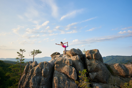 Young slim woman doing complicated yoga pose on lit by summer sun top of huge rock on green tree tops and clear blue sky background in the morning. Fitness and healthy lifestyle concept. Narajasanaの写真素材