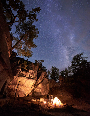 Campsite at night amid huge steep rock formation. Young tourist couple, man and woman standing near small tent lit by burning fire under clear dark starry sky. Tourism, traveling and nature concept.の写真素材