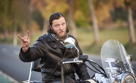 Young handsome bearded motorcyclist in black leather clothing sitting on shiny modern powerful motorbike showing horns sign on blurred vintage bokeh background of golden trees on bright sunny day.の写真素材