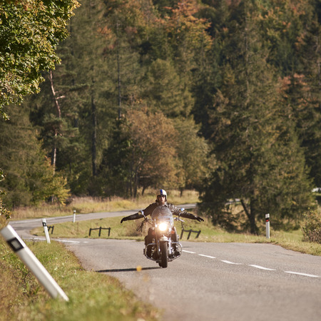 Bearded motorcyclist in helmet, sunglasses and black leather clothing riding motorbike with stretched out arms along empty asphalt road winding among tall green trees on bright sunny summer day.の写真素材