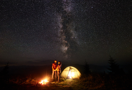 Romantic couple tourists boy and girl standing near illuminated tent by burning bonfire, holding hands under starry sky and Milky way, enjoying night camping in mountains. Tourism, traveling concept.の写真素材