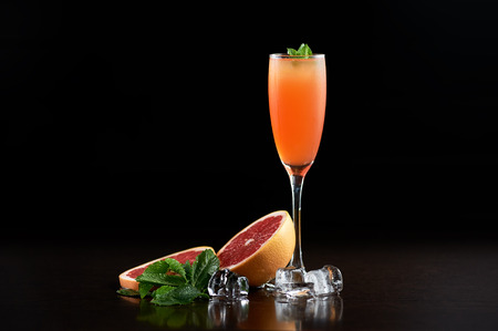 Tall narrow crystal cocktail glass with cold summer refreshing orange juicy drink, slices of grapefruit, fresh green mint leaves and transparent ice cubes on deep black copy space background.の写真素材