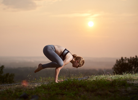 Profile of slim strong muscular girl doing handstand yoga exercises outdoors at dawn or twilight on copy space background of light pink summer sky and blurred misty landscape.の写真素材