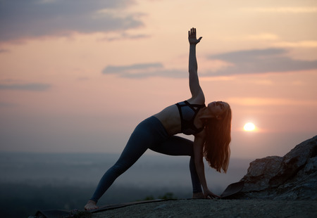 Silhouette of attractive long-haired young gymnast woman doing bending yoga exercises outdoors on high rocky hill on copy space background of cloudy beautiful pink sky and setting over horizon sun.の写真素材