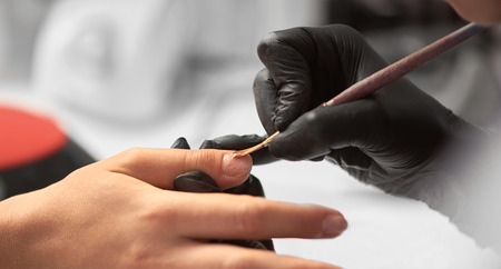 Close-up details shot of beautician hands in black rubber gloves applying with brush transparent gel polish on woman fingernail at beauty salon. Healthcare, beauty cosmetics, spa procedure concept.の写真素材