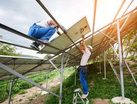 Solar photo voltaic panel system installing. Two technicians standing on ladder and platform top mounting heavy solar panel. Investment in alternative energy and professional construction concept.の写真素材