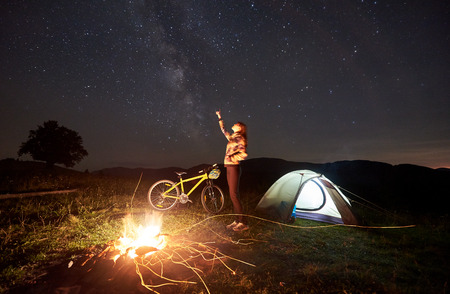 Young woman traveller resting at night camping, pointing at evening sky full of stars and Milky way, standing near burning campfire, illuminated tourist tent and mountain bike. Tourism conceptの写真素材
