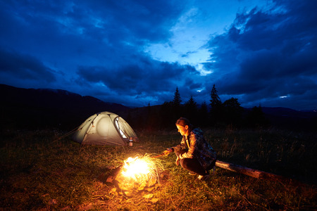 Active woman traveller having a rest at night camping in the mountains, sitting near burning campfire and illuminated tourist tent under beautiful evening cloudy sky. Tourism, outdoor activity conceptの写真素材