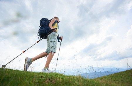 Low angle view of attractive woman hiker hiking in Carpathian mountain trail, walking on grassy hill, wearing backpack, using trekking sticks, enjoying summer day. Outdoor activity, lifestyle conceptの写真素材