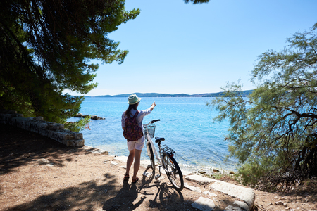 Back view of young woman in white closing, backpack and sunhat standing with bicycle on stony sidewalk under clear blue sky pointing at opposite shore on sparkling clear sea background on summer day.の写真素材