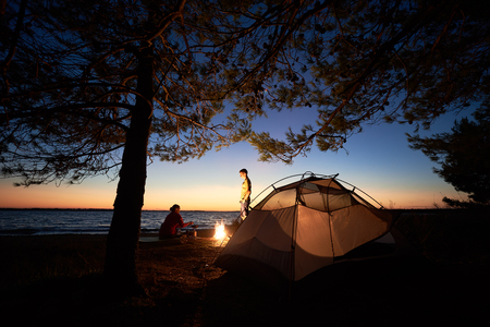 Camping on sea shore at night. Tourist tent under trees and young family, man and woman preparing food gas burner near campfire on blue evening sky and water background. Tourism and adventure concept.の写真素材