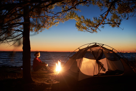 Night camping at sea coast under clear blue sky. Profile of smiling tourist woman sitting alone at campfire near tent on clear water and distant lights on horizon background. Tourism and recreation.の写真素材