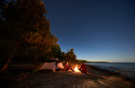Night camping on sea shore. Happy young man and woman hikers relaxing in front of tent at campfire under evening sky on clear blue water and green forest background. Active lifestyle concept.の写真素材