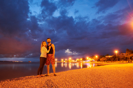 Young tourist couple, bearded man and long-haired woman standing embraced on resort town pebble beach, smiling to the camera at dusk on calm clear water and dramatic dark blue cloudy sky backgroundの写真素材