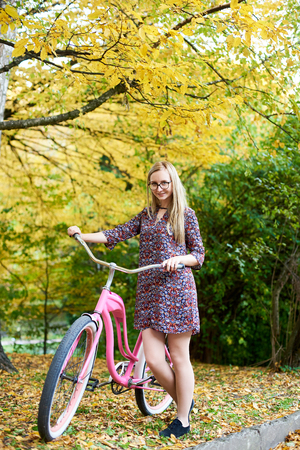 Slim happy blond fashionable attractive woman in glasses and short dress with pink lady bicycle standing alone under tall tree on beautiful green and golden autumn foliage sunny background.の写真素材