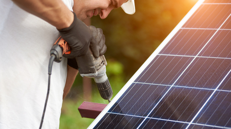 Close-up part view of professional worker in protective gloves connecting solar photo voltaic panel to metal platform using corded electrical screwdriver on blurred green summer background.の写真素材