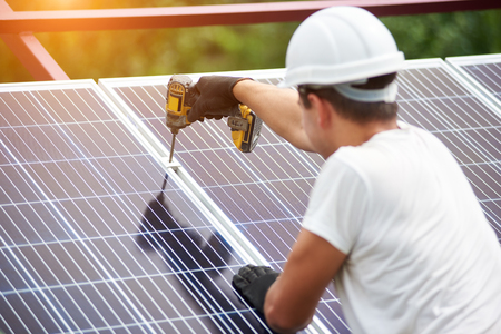 Back view of young technician in helmet connecting solar photo voltaic panel to metal platform using electrical screwdriver on shiny surface background. Stand-alone solar panel system installation.の写真素材