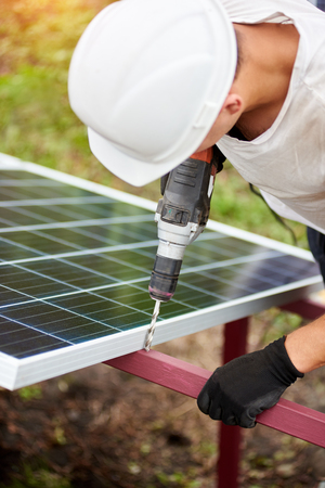 Close-up of professional technician working with screwdriver connecting solar photo voltaic panel to metal platform on warm sunny summer day. Stand-alone exterior solar panel system installation.の写真素材