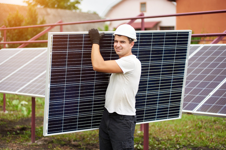 Smiling young worker in protective helmet carrying big shiny solar photo voltaic panel on exterior metal platform background on sunny summer warm day. Renewable ecological green energy concept.の写真素材