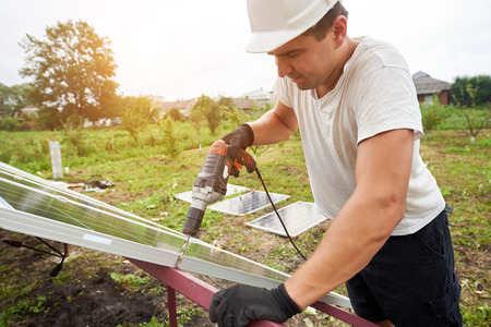 Young technician working with screwdriver connecting solar photo voltaic panel to metal platform on sunny green rural landscape background. Alternative sources, sun energy production concept.の写真素材