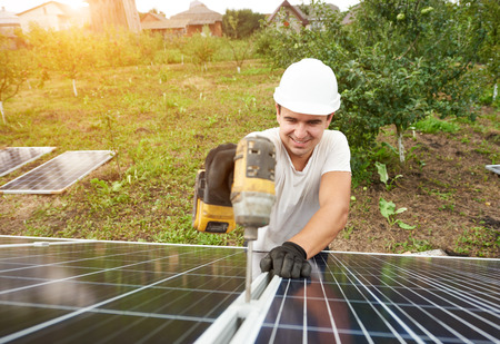 Smiling technician working with screwdriver connecting solar photo voltaic panel to exterior metal platform on sunny green orchard and rural houses background. Efficiency and professionalism concept.の写真素材