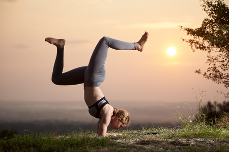 Profile of slim strong muscular blond girl doing difficult complicated yoga exercises outdoors at dawn on background of light pink summer sky and blurred misty horizon. Adho Mukha Vrksasanaの写真素材