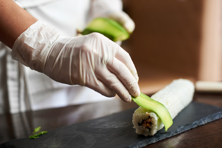 Close-up view of process of preparing rolling sushi at. Hand in glove decorates roll with sliced avocadoの写真素材