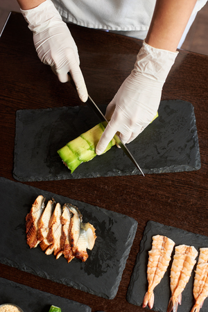 Closeup view of process of preparing rolling sushi. Chef is cutting roll on the black stone plate. View from the topの写真素材