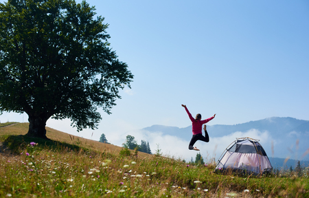 Back view of young happy woman jumping on blooming hill near small tourist tent and big tree under clear blue sky on bright summer morning on foggy mountains background. Tourism and traveling concept.の写真素材