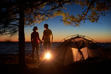 Night camping at lake. Silhouettes of tourist couple, boy and girl standing at campfire holding hands and looking at each other near tent. Tourism, happy relations and active lifestyle concept.の写真素材