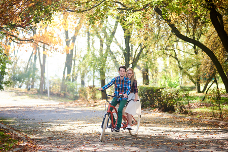 Young treveler couple, handsome bearded man and attractive blond long-haired woman cycling together tandem double bike by sunny alley with golden leaves on tall trees backgroundの写真素材