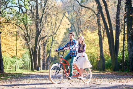 Young tourist couple, handsome bearded man and attractive blond long-haired woman in glasses cycling together tandem double red bike by sunny alley with golden leaves on tall trees backgroundの写真素材