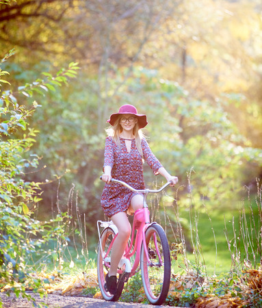 Slim happy smiling blond fashionable attractive girl in glasses, short dress and pink hat riding lady bicycle along paved park alley on beautiful green and golden trees lit by bright sun background.の写真素材