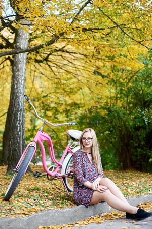 Blond attractive woman in glasses and fashionable short dress sitting alone on ground at pink lady bicycle under tall tree with golden leaves on beautiful colorful autumn foliage evening background.の写真素材