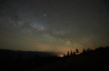 Fantastic mountains view at night. Steep hills covered with forest, fir-trees silhouettes against dark blue sky with bright sparkling stars and distant light on horizon. Beauty of nature concept.の写真素材