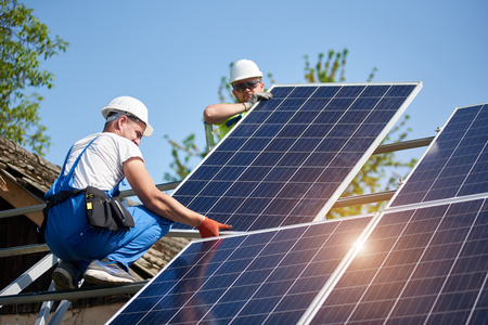 Two professional technicians adjusting heavy solar photo voltaic panel to metal platform on blue sky background. Stand-alone solar panel system installation, efficiency and professionalism concept.の写真素材