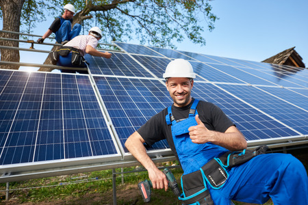 Portrait of smiling technician with electrical screwdriver showing thumb-up in front of unfinished high exterior solar panel photo voltaic system with team of workers on high platform.の写真素材