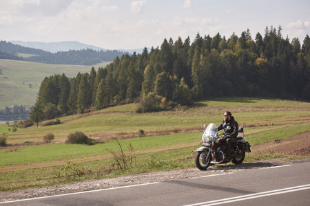 Handsome bearded biker in black leather jacket sitting on cruiser motorcycle on roadside on blurred background of empty twisty road with white marking on sunny summer day.の写真素材