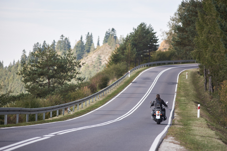 Back view of motorcyclist in black leather outfit with long hair riding cruiser motorbike up twisted empty asphalt road on bright sunny summer day by green forest trees under clear blue cloudless sky.の写真素材