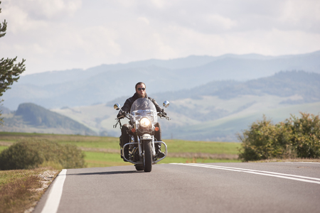 Alone motorcyclist in black leather outfit riding motorbike along twisted road on bright sunny summer day on background foggy distant green woody hills under blue morning sky.の写真素材