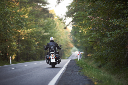 Back view of motorcyclist in black leather jacket and white helmet riding motorbike along hilly road between tall green trees on sunny summer day. Active lifestyle, love to adventures concept.の写真素材
