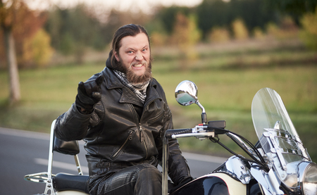 Handsome bearded motorcyclist in black leather clothing sitting on shiny powerful cruiser motorbike showing horns sign on blurred vintage bokeh background of golden autumn trees.の写真素材